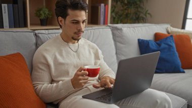 Genç İspanyol adam rahat oturma odasında dizüstü bilgisayarı ve kahveyle kanepede dinleniyor, modern ev yaşam tarzını sergiliyor..