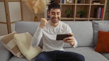 İspanyol adam elinde akıllı telefonuyla evde kutlama yapıyor, evdeki koltukta oturuyor, modern bir oturma odasında yumruğunu kaldırarak mutluluğu gösteriyor..