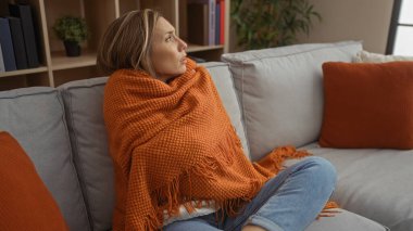 Portakal rengi battaniyeye sarılı, modern dekorlu, yumuşak yastıklı bir kadın..