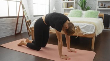 Konforlu yatak odası ortamında yoga yapan bir kadın yeşil yastıklı bir yatağın yanında pembe bir paspas üzerinde geriniyor ve büyük bir pencereden gelen doğal ışıkla dans ediyor..