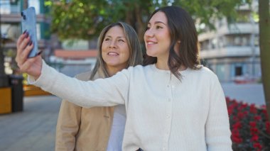 Anne ve kızı şehir parkında selfie çekiyorlar güneşli bir günde açık havada aile sevgisini ve neşesini sergiliyorlar.