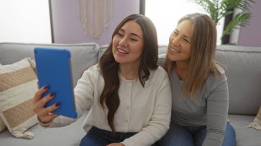 Anne ve kızı oturma odasında selfie çekiyorlar, aile bağı ve mutluluğu sergiliyorlar, evde birlikte oturan kadınlar var..