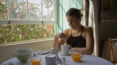 Etrafı aydınlık ve yeşilliklerle çevriliyken rahat bir restoranda kahvesini yudumlayan genç bir kadın, huzur dolu bir kahvaltı sahnesini işaret ediyor.