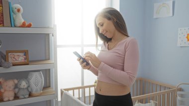 Akıllı telefon kullanan bir kadın beşiği olan rahat bir yatak odasında duruyor. Etrafı yumuşak oyuncaklarla çevrili ve pencerelerden gelen hafif bir ışık, evin içinde huzurlu bir atmosfer yaratıyor..