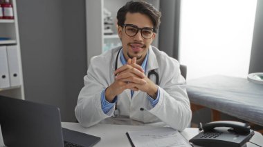 Gözlüklü ve steteskoplu laboratuvar önlüklü genç adam modern klinikte güvenle gülümsüyor. Laptop ve telefonla sağlık sektöründe profesyonellik gösteriyor.