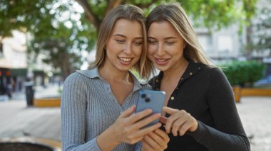Şehir parkında akıllı bir telefona bakarken gülümseyen ikizler, doğal yeşillik dolu bir şehir ortamında aile bağı ve mutluluğu gözler önüne seriyorlar..