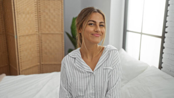 Женщина улыбается в постели с полосатой пижамой в яркой, уютной спальне, демонстрируя расслабленную утреннюю атмосферу с естественным светом, струящимся через окно.