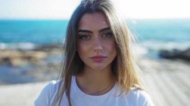 Sahilde güneşin altında, kumsalda, kapalı gözlerle beyaz gömlekli, açık mavi gökyüzü ve deniz manzarasını izleyen bir kadın..