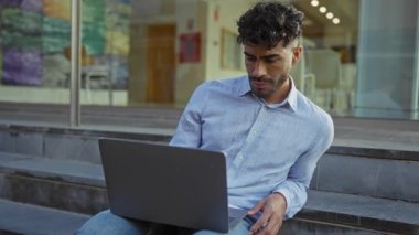 Kentsel basamaklarda oturan sakallı genç bir adam, dizüstü bilgisayarla stresli görünüyor, şehir merkezinde açık havada, baş ağrısı ya da konsantrasyon sorunu öneriyor..