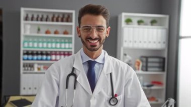 Laboratuvar önlüğü giymiş bir hastane ortamında genç bir adam elleriyle bir kalp oluşturuyor. Kliniğin iç kısımlarında güvenle gülümsüyor..