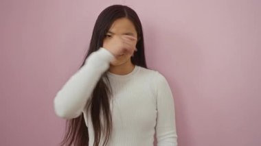 Uzun saçlı bir kadın, beyaz bir süveter içinde yumuşak pembe bir arka plana karşı güven içinde poz veriyor..