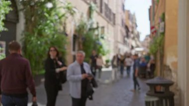Bulanık sokak sahnesinde, Roma, İtalya 'da tarihi bir kaldırım taşı caddesinin üzerinde yürüyen ve sosyalleşen insanlar görülüyor..