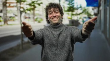 Genç adam, güneşli bir günde, şehir caddesinde gülümseyip, açık havada neşe ve güveni ifade ederek kendine sarılır..