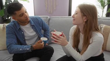 Genç çift, rahat bir oturma odasında rahat bir kanepede birlikte kahve içiyorlar. Yumuşak ışıklandırmalı rahat ve samimi bir ev atmosferi öneriyorlar..