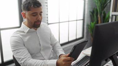 Genç adam, modern işyerindeki bilgisayarla çalışırken tablet kullanarak ofiste oturuyor ve iç mekan ortamını vurguluyor..