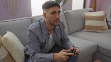 Genç adam rahat bir oturma odasındaki kanepede video oyunları oynuyor, odak ve konsantrasyon gösteriyor, modern dekorasyonla sıradan bir iç mekan görüntüsü yakalıyor..
