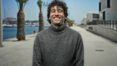 Sıcak bir süveter içinde gülümseyen bir adam, palmiye ağaçları ve modern mimariyle çevrili, güneşli açık havada sıcak ve rahat bir cazibe yayıyor..