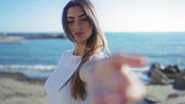 Deniz kenarında el kol hareketi yapan bir kadın, açık deniz gününde okyanus manzaralı güneşli gezintiye davet ediyor, güzel, zarif ve kendine güvenen bir kadın..