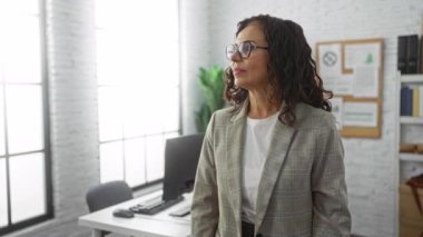 Gözlüklü ve ceketli kendine güvenen İspanyol kadın modern bir ofiste kolları çapraz duruyor. Parlak pencereleri ve dekorasyonu var. Profesyonelliği ve liderliği aktarıyor..