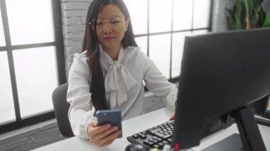 Gözlüklü kadın, modern ofiste masasında akıllı telefon kullanıyor. Büyük pencereleri ve bilgisayar düzeneği var..