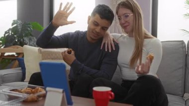 Görüntülü konuşma sırasında yemek yemekten hoşlanan çift oturma odasında tabletle birlikte el kol hareketleri yapıyor.