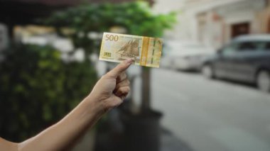 Norveçli 500 kronu arka planda arabaları olan bir şehir caddesinde, şehir merkezinde Norveç paralarını gösteren bir banknotta tutuyorlar..