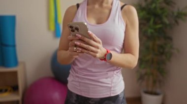 Pembe bluz giyen bir kadın spor salonunda spor aletleri ve arka planda görülebilen spor aletleriyle akıllı telefon kullanıyor. Bu da modern aktif yaşam tarzını gösteriyor..