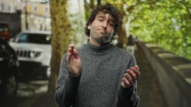 Arka planda ağaçlar ve arabalarla çevrili gri bir süveter giyen Latin asıllı genç adam, bir şehir caddesinde ciddi şekilde alkışlıyor..