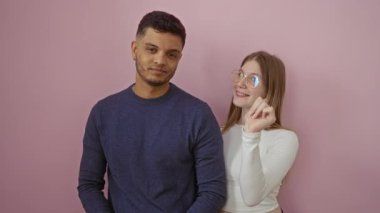 Genç bir çift pembe bir duvar arkasına başparmağıyla gülümsüyor, pozitif ve dostane bir ilişki dinamiği sergiliyorlar..