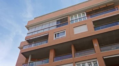 Açık mavi gökyüzü altında kırmızı tuğlalı ve balkonları olan modern apartman binası canlı bir şehir ortamında kentsel mimari ve yaşam tarzını gözler önüne seriyor..