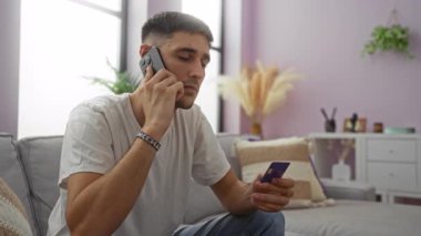 Oturma odasında elinde kredi kartı ve akıllı telefon olan genç adam sohbete odaklanmış, dekorla çevrili modern koltukta oturuyor, günlük ev dekorunu vurguluyor..
