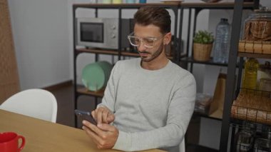 Modern ofis mutfağında akıllı telefon kullanan, minimalist dekorlar ve gerekli aletlerle çevrili, sıradan ve odaklanmış tavırlar sergileyen genç bir adam..