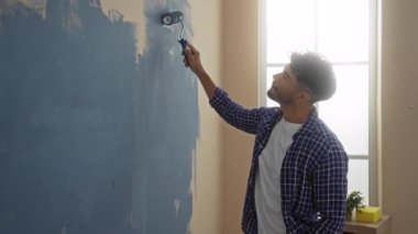 Genç adam modern evin iç kısımlarında duvar boyuyor. Pencereden süzülen ışık huzurlu bir atmosfer yaratıyor.