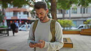 Şehir parkında telefon kullanan genç bir adam. Sırt çantası ve günlük süveter ile şehir hayatı ile çevrili.