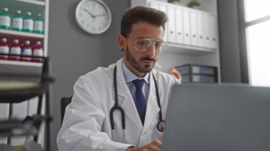 Doktor klinikte dizüstü bilgisayarla çalışırken, stresi ifade ederken, tıbbi malzemeler ve profesyonel dekor ile çevriliyken, saat arka planda zaman gösteriyor..