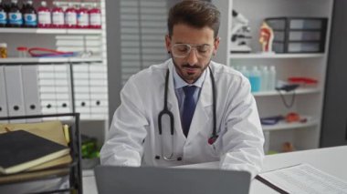 Ciddi genç İspanyol doktor klinikte gözlük çıkartıyor, dizüstü bilgisayara odaklanmış, stetoskopla, profesyonel tıbbi atmosferi somutlaştırıyor..