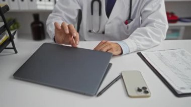 Steteskoplu beyaz önlüklü doktor, tıbbi malzeme ve belgelerle çevrili bir klinikte, profesyonel sağlık hizmetlerini gösteren bir dizüstü bilgisayar kullanıyor..