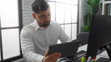 Ofisteki genç adam, bilgisayar başında odaklanmış bir ifadeyle çalışıyor, gözlük takıyor ve beyaz bir gömlek giyiyor, büyük pencerelerden gelen modern bir iç mekan ve doğal ışık ile çevrili..