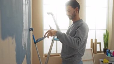 Ev dekoru ve doğal ışık ile çevrili telefon ve silindir tutarken parlak oturma odasındaki duvarı boyayan genç adam.