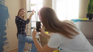 Evini yenileyen kadınlar, biri diğerinin merdiven üzerinde resim çekerken elinde boya silindiri, etrafı kutular ve malzemelerle çevrili, kapalı alanda neşeli anlar yakalıyorlar..