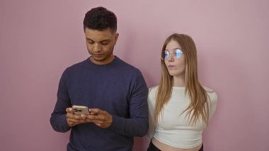 Pembe bir duvarın karşısında duran bir adamın telefonunu kullanan bir kadın, modern bir ilişki ortamında merak ve şüphe uyandırır..