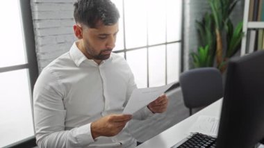 Genç adam modern ofiste büyük pencerelerden yansıyan doğal ışık ile belge okuyor ve odaklanmış iş için profesyonel bir ortam yaratıyor.