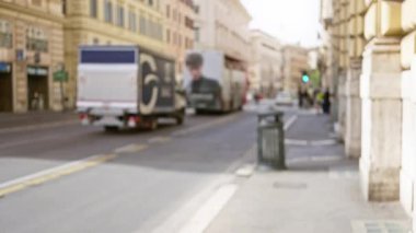 Arka planda tarihi binalar ve araçlarla güneşli bir Roma caddesinde yürüyen bir adamın bulanık görüntüsü tipik İtalyan şehir yaşamını gözler önüne seriyor..