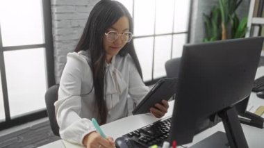 Tablet kullanan ve yanında bilgisayar olan bir ofiste notlar alan bir kadın, iç mekan bitkileri ve parlak pencerelerle çevrili, modern bir işyeri ayarını yakalıyor..