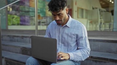 Genç İspanyol bir adam şehir ortamında dizüstü bilgisayar kullanıyor, mavi bir gömlek giyiyor, güneşli bir günde merdivenlerde oturmuş, modern yaşam tarzı ve teknolojisini aktarıyor..
