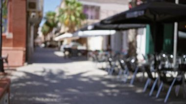 Avrupai arka planlarının bulanık olduğu bir Avrupa caddesinin olduğu açık hava manzarası. Palmiye ağaçları ve şemsiyelerin altında açık kafe oturma masaları ve sandalyeler de dahil.