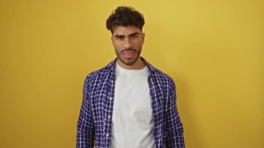 Sakallı genç adam, orta parmaklarıyla canlı sarı bir arka plana karşı bir jest yapıyor. Kareli gömlek giyiyor, meydan okuma ve tavır sergiliyor..