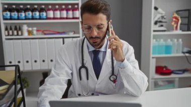 Klinikte beyaz önlüklü genç adam, teşhis ekipmanları ve tıbbi gereçlerle telefonda konuşuyor, profesyonel ve modern bir sağlık hizmeti sunuyor..