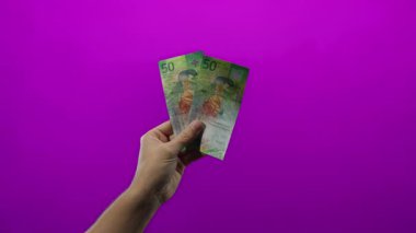 İsviçre frangı banknotları tutan adam canlı mor bir duvara karşı, modern bir bağlamda finans ve para yönetimi vurguluyor.