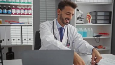 Tıbbi malzeme ve ekipmanlarla dolu bir klinikte kadehleri ayarlarken gülümseyen genç İspanyol erkek doktor profesyonellik ve güven yayıyor..